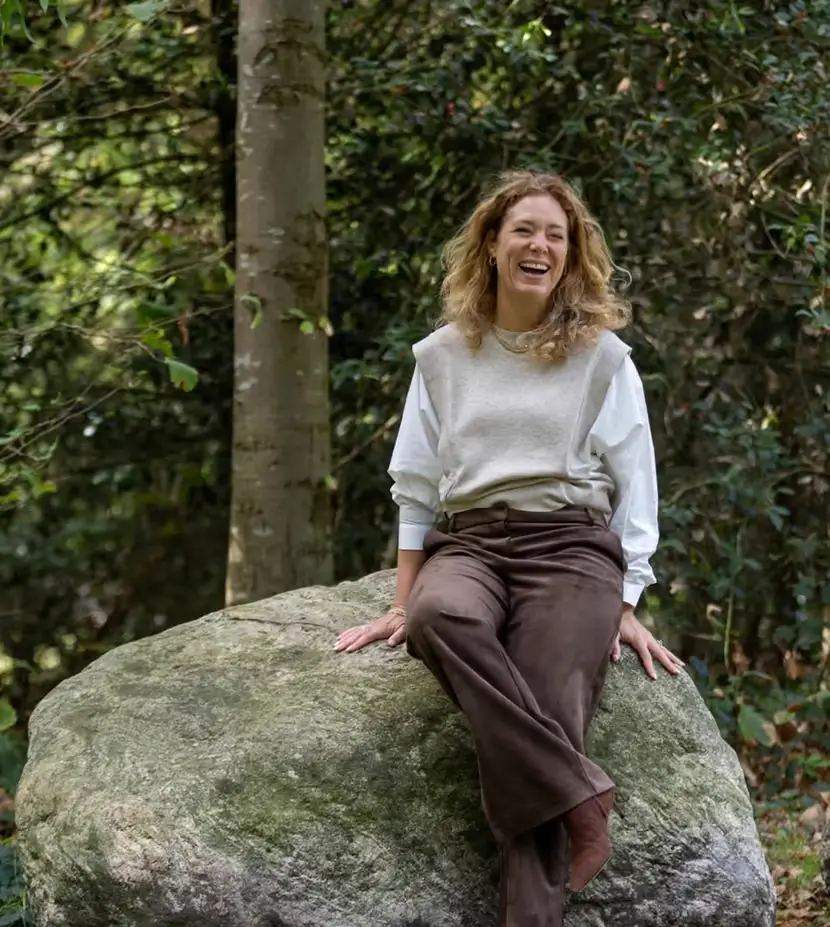 Suzanne van Hulten lachend op een grote kei in een bosrijke tuin